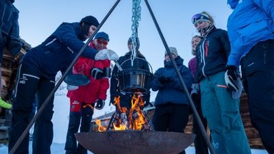 Eine Gruppe von Menschen steht im Schnee um ein Lagerfeuer mit einem hängenden Kessel.