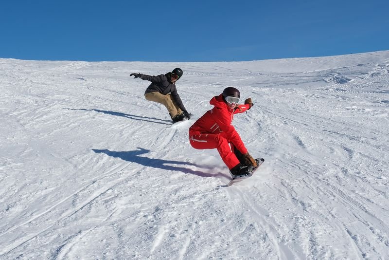 Two snowboarders in helmets and goggles ride down a snowy mountain slope under a clear sky.