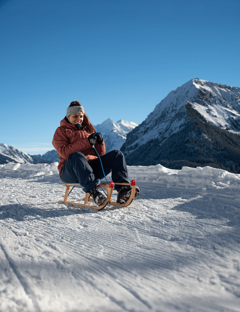 Person sitting on a sled in snowy mountains, wearing a jacket and headband, with clear blue sky.