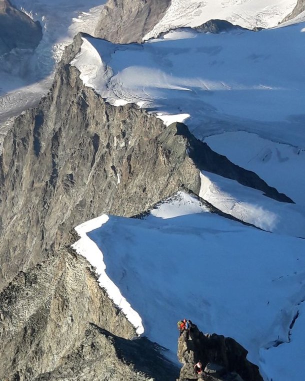 Climbers with helmets and harnesses ascend a rocky ridge in a snowy mountain landscape.