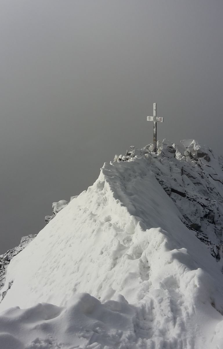 Snow-covered mountain peak with a cross, surrounded by foggy sky.