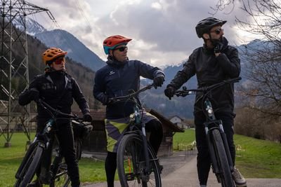 Three cyclists with helmets and sunglasses on mountain bikes in a rural area with mountains in view.