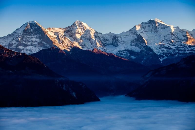 View of snow-covered Alpine mountains at sunrise, with fog in the valley below.