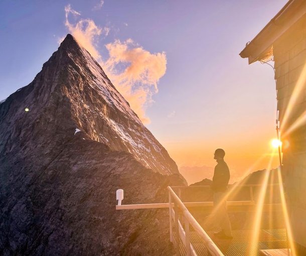 Person standing on a balcony at sunset, overlooking a mountain peak with snow patches.