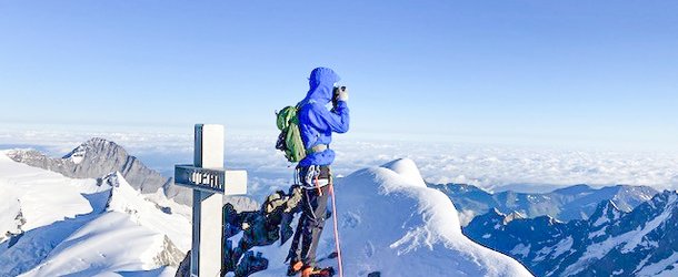 Climber in blue jacket on snowy peak with helmet, harness, and rope, near a cross, overlooking mountains.