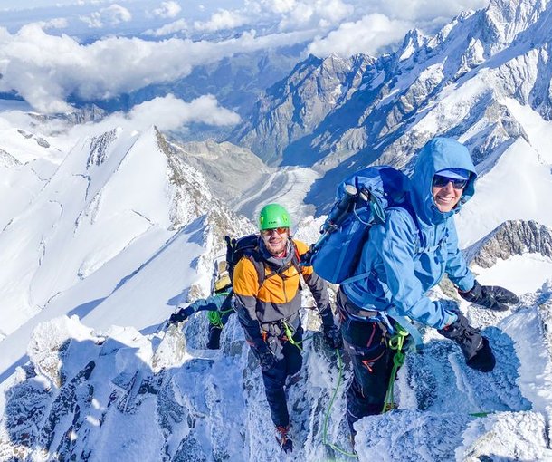 Two climbers with helmets and harnesses ascend a snowy mountain ridge with a scenic alpine backdrop.