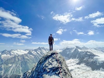 Person auf Berggipfel mit Kletterausrüstung und Seil, umgeben von verschneiten Alpen.