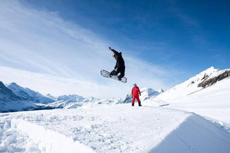 Snowboarder in mid-air jump on snowy slope, with another person in red suit watching, mountains in background.