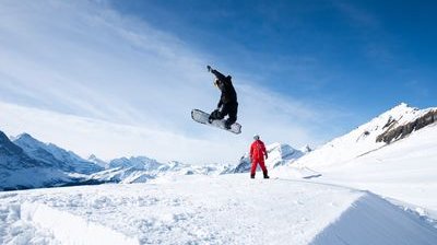 Snowboarder in der Luft bei einem Sprung auf schneebedecktem Hang, mit einer anderen Person in einem roten Anzug, der zuschaut, Berge im Hintergrund.