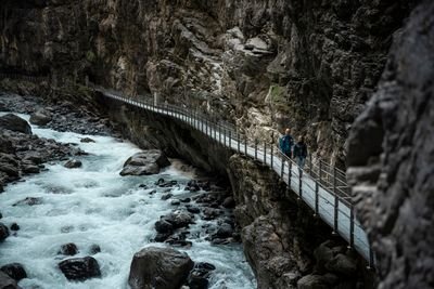 Zwei Personen mit Rucksäcken und Helmen wandern auf einem schmalen Steg entlang einer Felswand über einem reißenden Fluss.