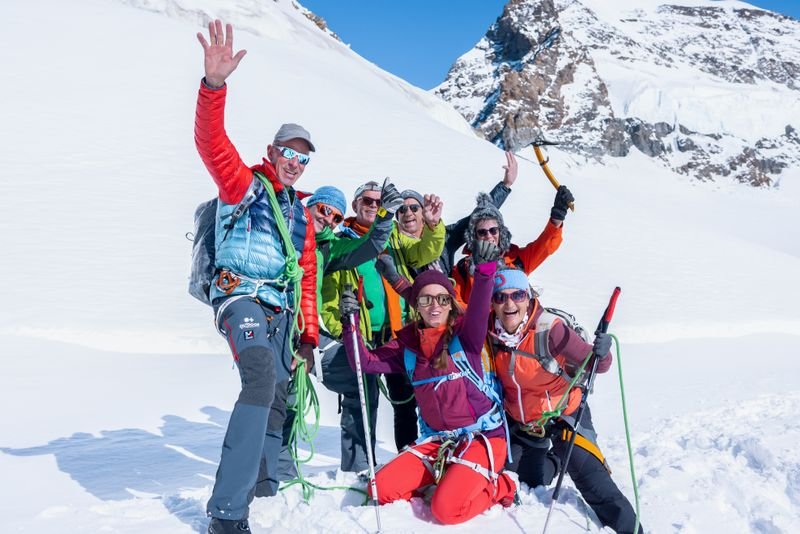 A group of people with climbing equipment and helmets is standing in the snow in front of a mountain backdrop.