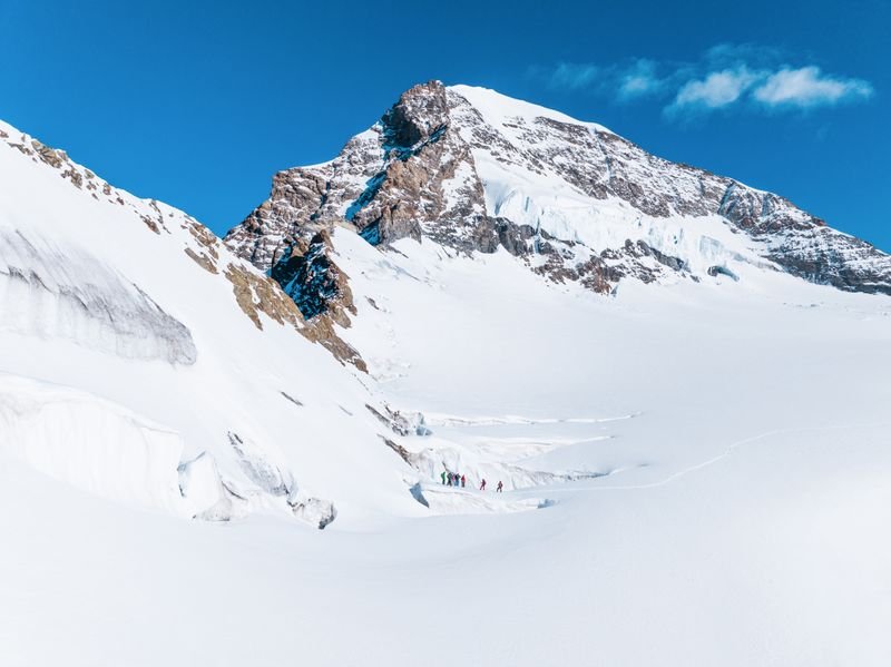 People with winter equipment are hiking on a snow-covered mountain glacier under a blue sky.
