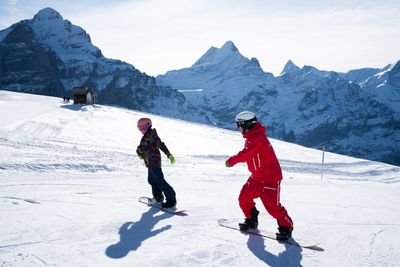 Two snowboarders in helmets and winter gear on a snowy mountain slope with distant peaks.