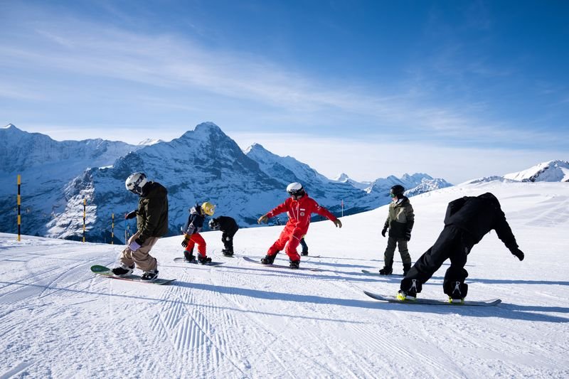 Snowboarders with helmets and winter gear on a snowy mountain slope with distant peaks.