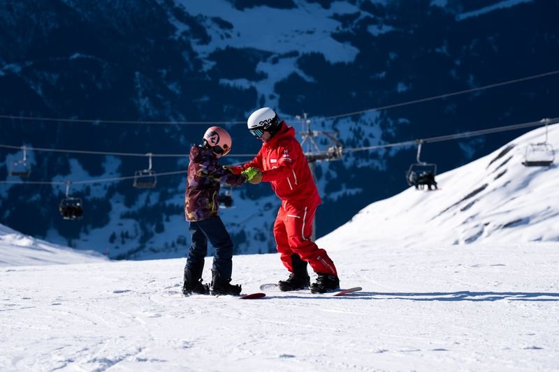 Two people snowboarding on a snowy mountain, wearing helmets and winter gear, with ski lifts behind.