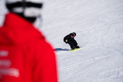 A person snowboards down a snowy slope, wearing a helmet and goggles, with a guide in a red jacket nearby.
