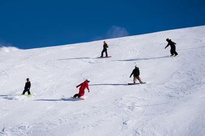 Five people snowboarding downhill on a snowy mountain, wearing helmets and winter gear.