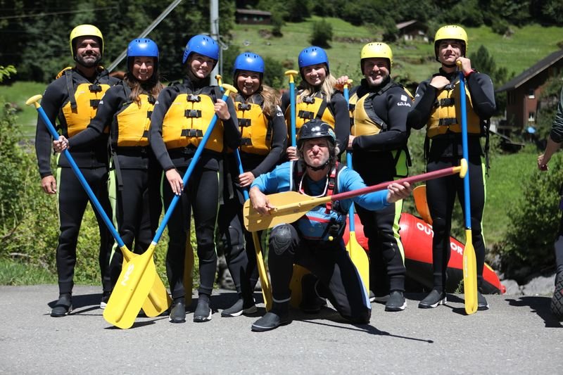 A group in wetsuits, life jackets, and helmets holding paddles; in the background mountains.
