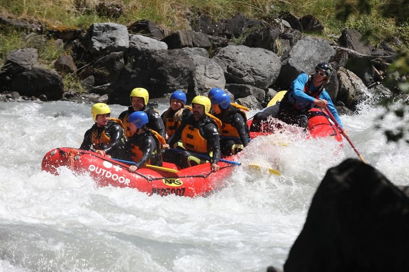 Group rafting in white water in a red inflatable boat, wearing helmets and life jackets on a river.