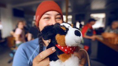 Person hält ein Plüschtier in einer Hütte, trägt eine rote Mütze. Hintergrund unscharf.