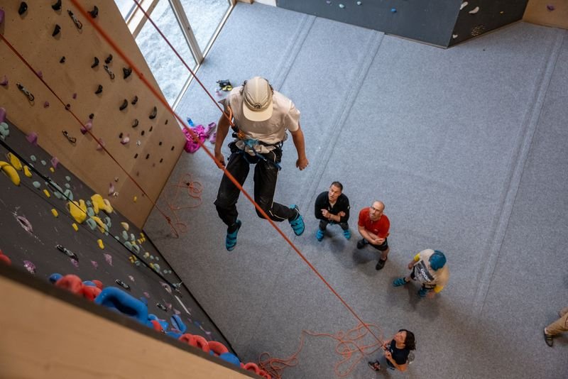 Person in climbing gear rappelling indoors, with others watching below on a padded floor.