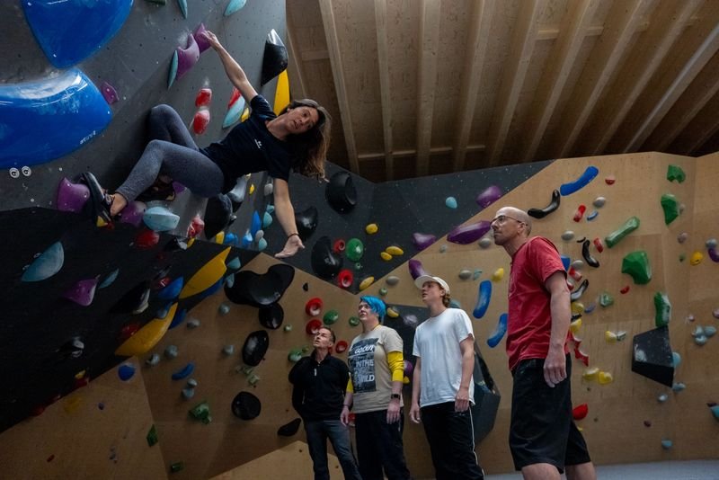 Person klettert an einer Indoor-Boulderwand mit bunten Griffen und wird von vier anderen in legerer Kleidung beobachtet.