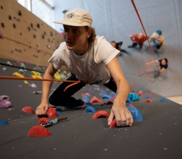 Person climbing indoor wall with colorful holds, wearing a white shirt, cap, and harness with rope.