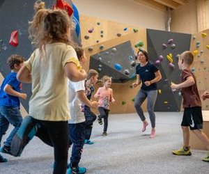 Children and an instructor in a climbing gym, wearing climbing shoes, with a bouldering wall in view.