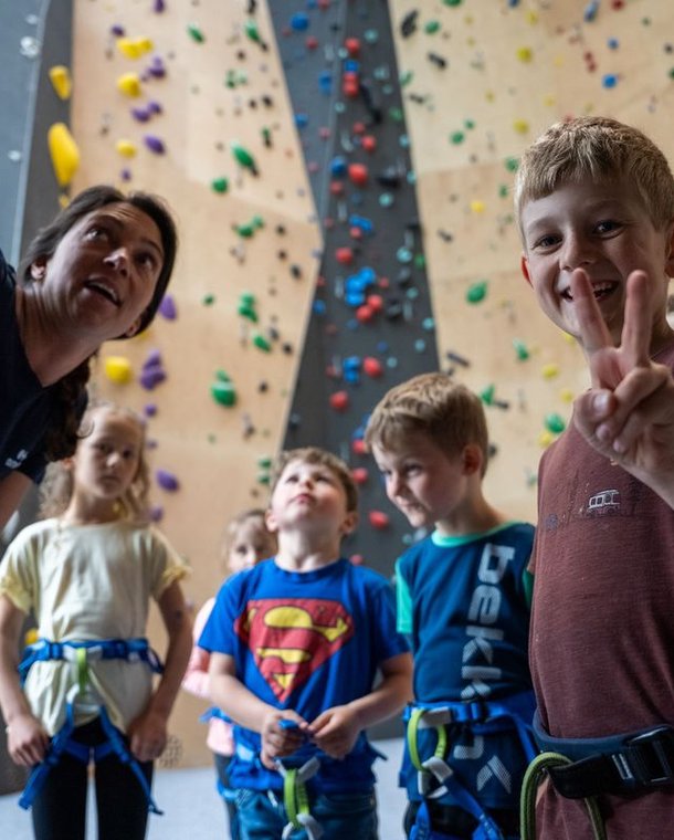 Kinder in Gurten hören einem Trainer an einer Indoor-Kletterwand zu.