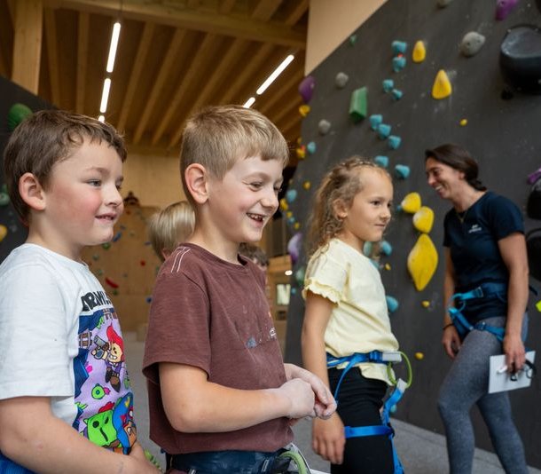 Kinder mit Gurten in einer Indoor-Kletterhalle, bunte Griffe an den Wänden, erwachsener Betreuer anwesend.