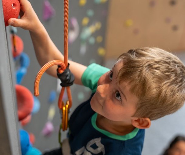 Child climbing indoor wall with harness and rope, colorful holds visible, adult in background.