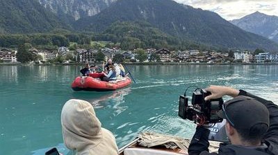 People in an inflatable boat on a lake, surrounded by mountains, with camera shots from the boat.