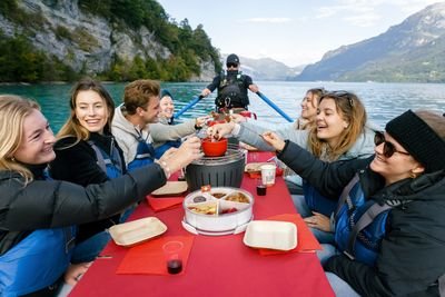 People in life jackets are eating fondue on a boat on a lake, surrounded by mountains.