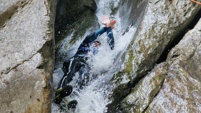 Person rutscht in einem felsigen Canyon im Wasser, trägt einen Neoprenanzug und einen Helm; zwei Guides beobachten.
