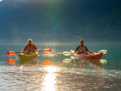 Zwei Personen mit Rettungswesten und Hüten, die auf einem ruhigen See mit Bergen im Hintergrund Kajak fahren.