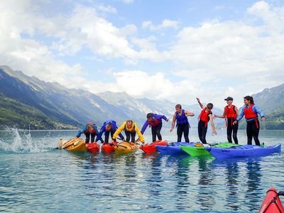 Sieben Personen in Rettungswesten balancieren und springen auf Kajaks auf einem See mit Bergen im Hintergrund.