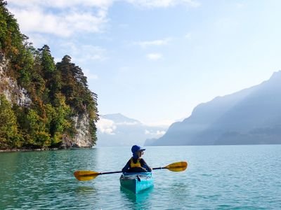 Person in blauem Kajak mit Paddel auf einem See, umgeben von Bergen und bewaldeten Klippen.