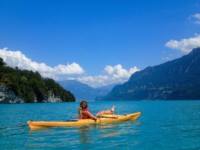 Person in gelbem Kajak auf blauem See, umgeben von Bergen und bewaldeten Ufern, unter blauem Himmel.
