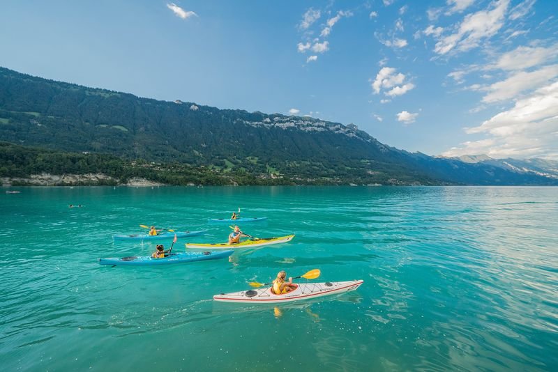 Personen in Kajaks paddeln auf einem türkisfarbenen See, umgeben von Bergen und blauem Himmel.