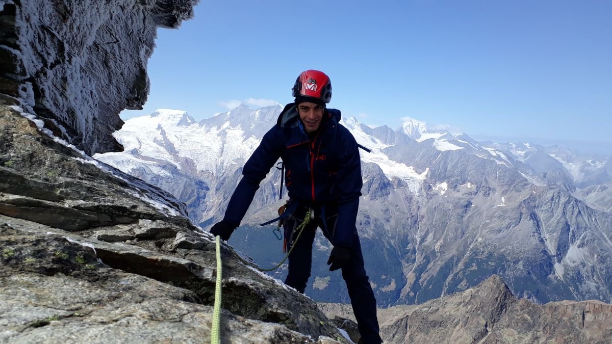 Climber with helmet and harness scaling a rocky mountain with snowy peaks in the background.