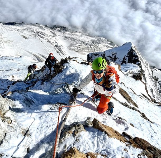Climbers with helmets and harnesses ascend a snowy mountain ridge, using ropes for support.