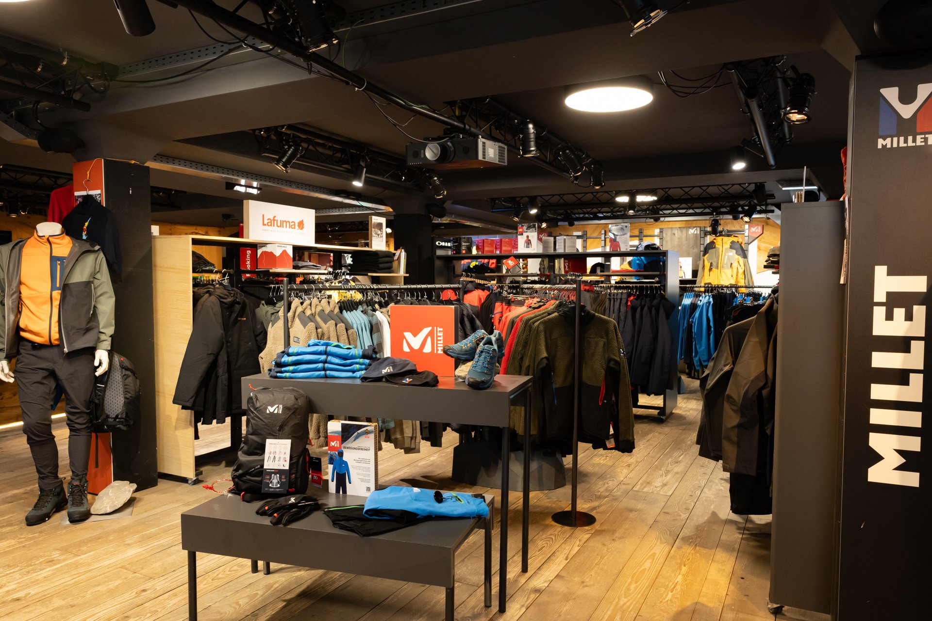Innenansicht eines Outdoor-Geschäfts mit Jacken, Hosen und Mützen auf Kleiderständern und Tischen.