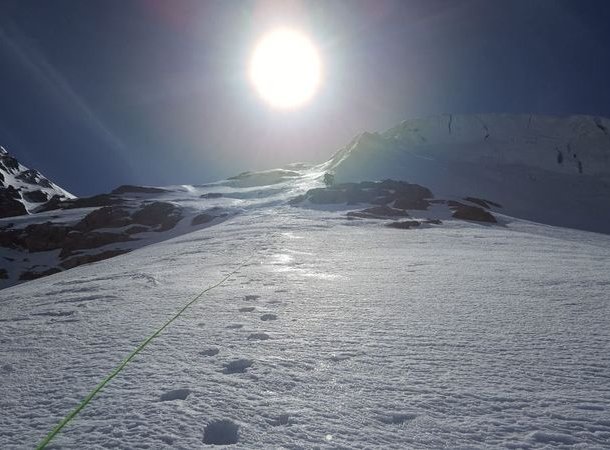 Snowy mountain slope with a visible climbing rope and footprints under a bright sun.