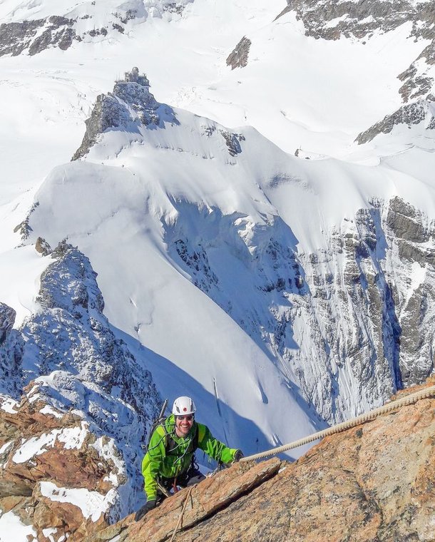 Climber in green jacket with helmet and rope ascends snowy mountain ridge with peaks in background.