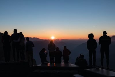 People are standing on a mountain plateau at sunset, silhouetted against a mountain landscape.
