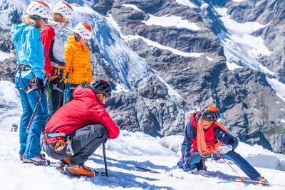 People in climbing gear with helmets and ropes on a snow-covered mountain in front of a rocky backdrop.