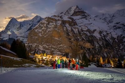 A group of people with sleds and winter gear on a snowy mountain slope at dusk, with peaks behind.