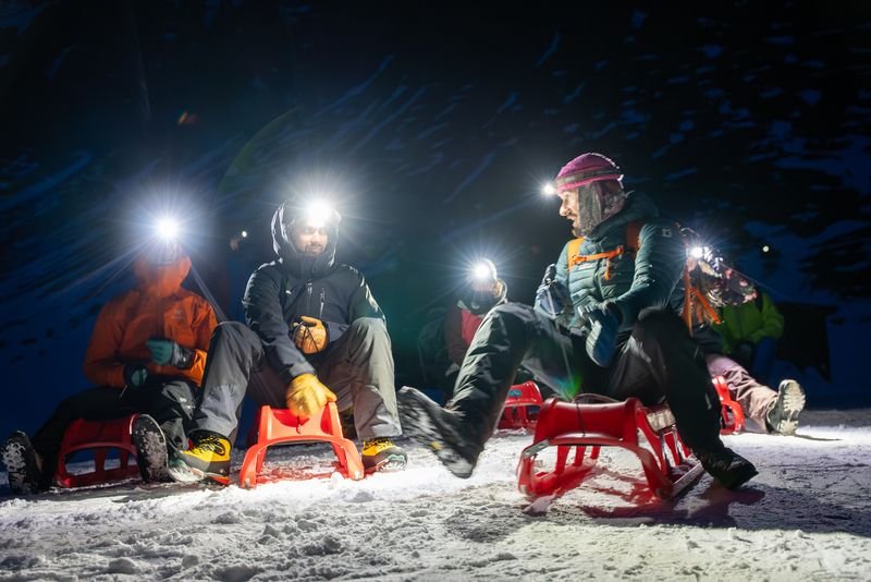 People on sleds wearing headlamps and winter gear, sitting on snow in a mountainous area at night.