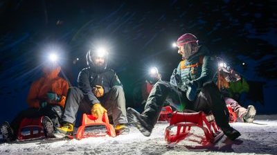 Menschen auf Schlitten mit Stirnlampen und Winterkleidung, die nachts auf Schnee in einem bergigen Gebiet sitzen.