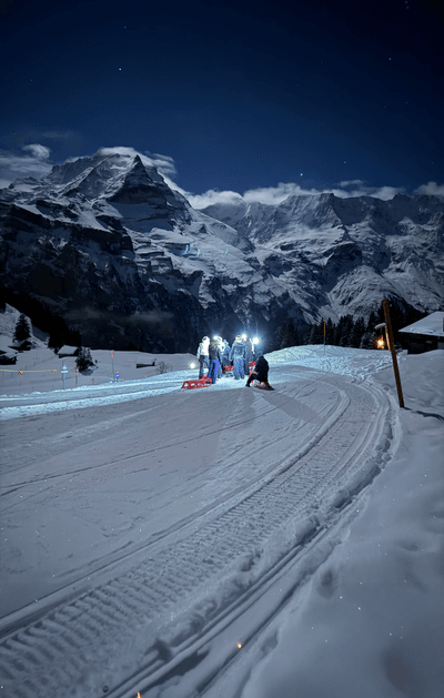 People with sleds and headlamps on a snowy mountain path at night, with mountains in the background.
